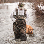 Man in camouflage outfit and hat, accompanied by a dog, standing in water and wearing AquaWade Pro Insulated Duck Waterfowl Hunting Chest Waders.