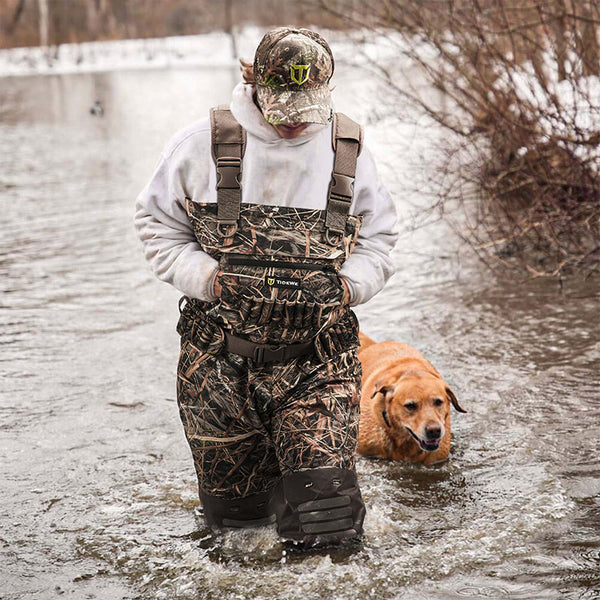 Man in camouflage outfit and hat, accompanied by a dog, standing in water and wearing AquaWade Pro Insulated Duck Waterfowl Hunting Chest Waders.