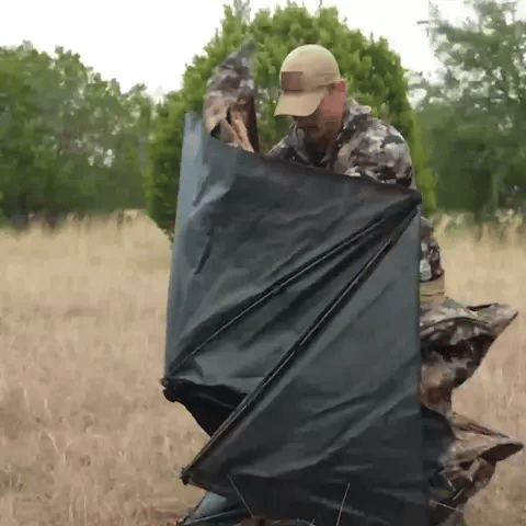 Man in camouflage holding the Occult See Through Hunting Blind 2-3 Person Pop Up Ground Deer Blind, showcasing its portable design and outdoor usability.