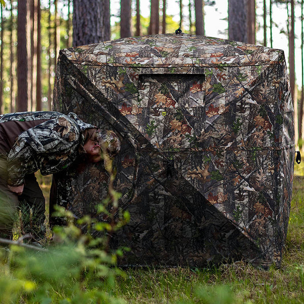 Person setting up the Occult See Through Hunting Blind 2-3 Person Pop Up Ground Deer Blind in a forested area, showcasing its camouflage design and spacious interior.