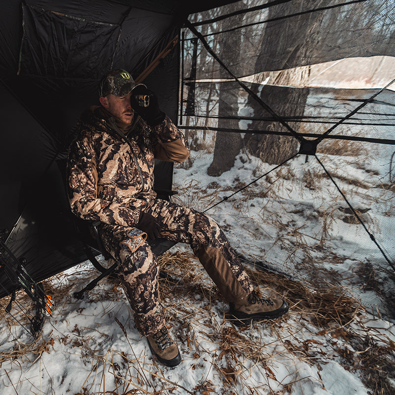 A man sits in a tent using a telescope, demonstrating the 270 Degree 3 Full Panels See Through Hunting Blind 2 Sets Bundle.