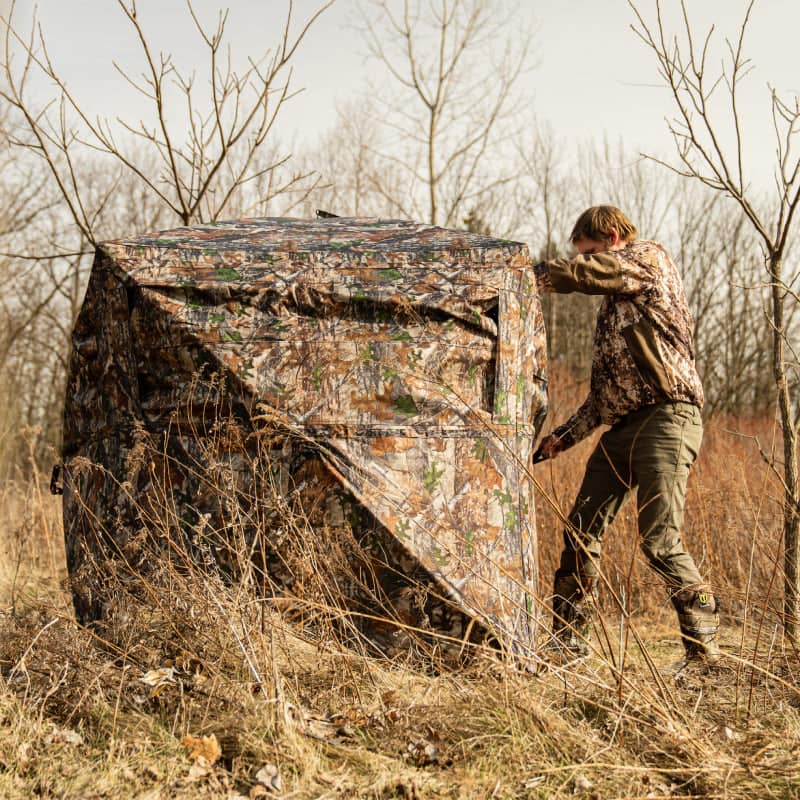 Man setting up the Occult See Through Hunting Blind 2-3 Person Pop Up Ground Deer Blind in a grassy field.