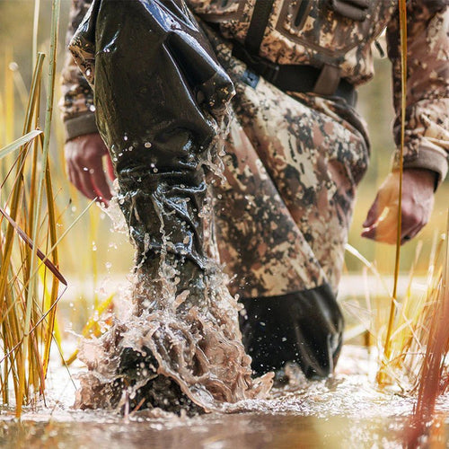 A person in camouflage DeepWade Zipped Duck Waterfowl Hunting Breathable Chest Waders stands in a muddy puddle, showcasing durable, waterproof, and insulated features.